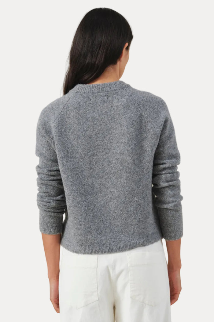 A woman viewed from the back wearing a heather gray long-sleeve knit sweater and white pants, standing against a plain light beige studio background.