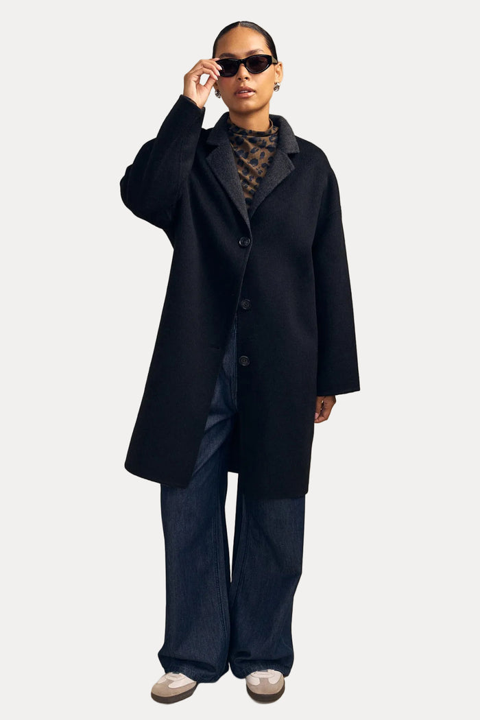 A model wearing a long black wool-blend coat over a patterned top and wide-leg blue jeans, standing against a solid beige studio background.
