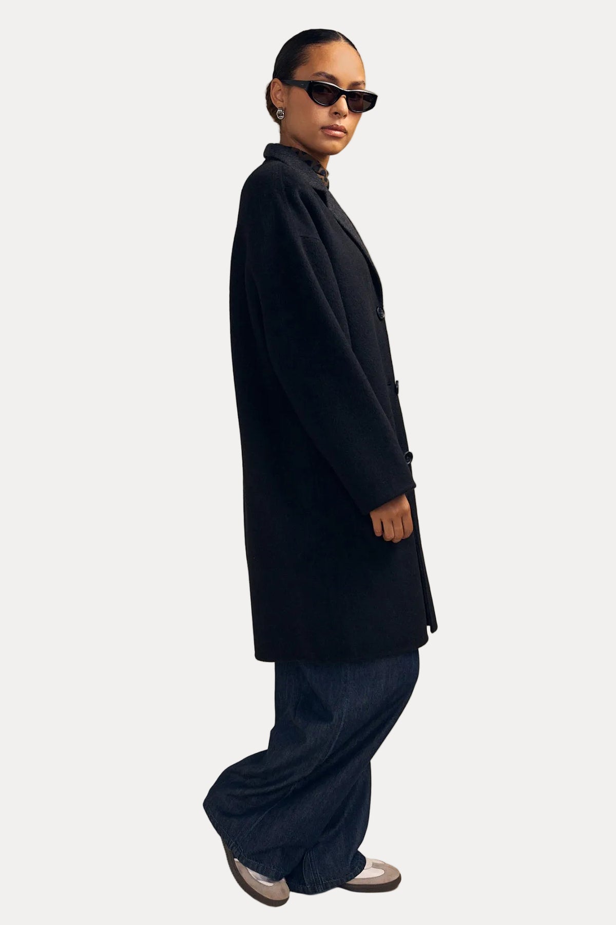 A model wearing a long black coat, wide-leg dark blue jeans, black sunglasses, and silver sneakers, standing in profile against a solid beige studio background.