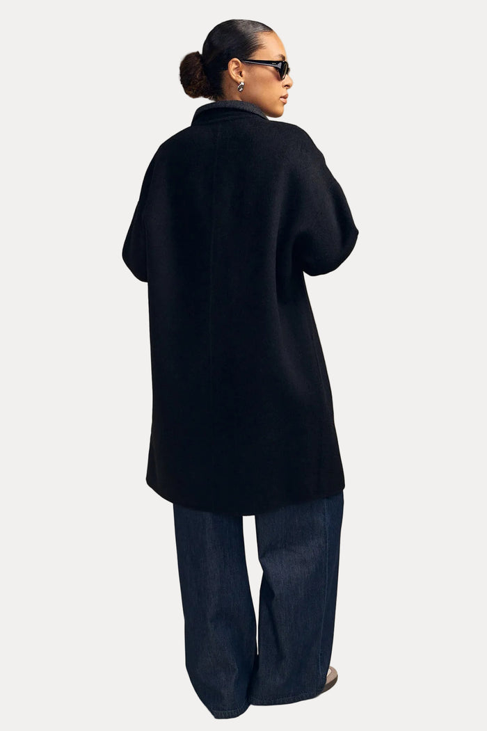 A model shown from the back wearing a black oversized coat and dark wide-leg jeans, standing against a solid beige studio background.