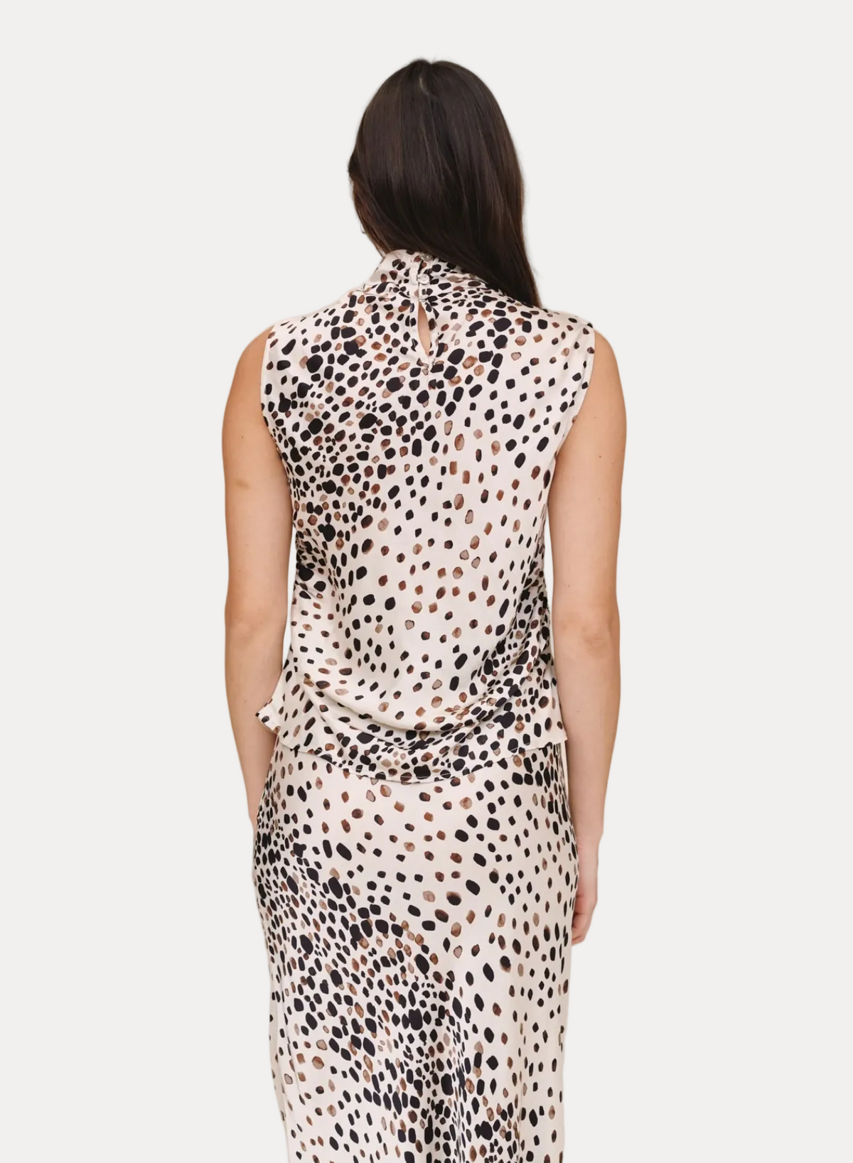 A woman viewed from the back wearing a sleeveless high-neck dress with a black, brown, and beige spotted animal print on a cream background, set against a plain beige studio backdrop.