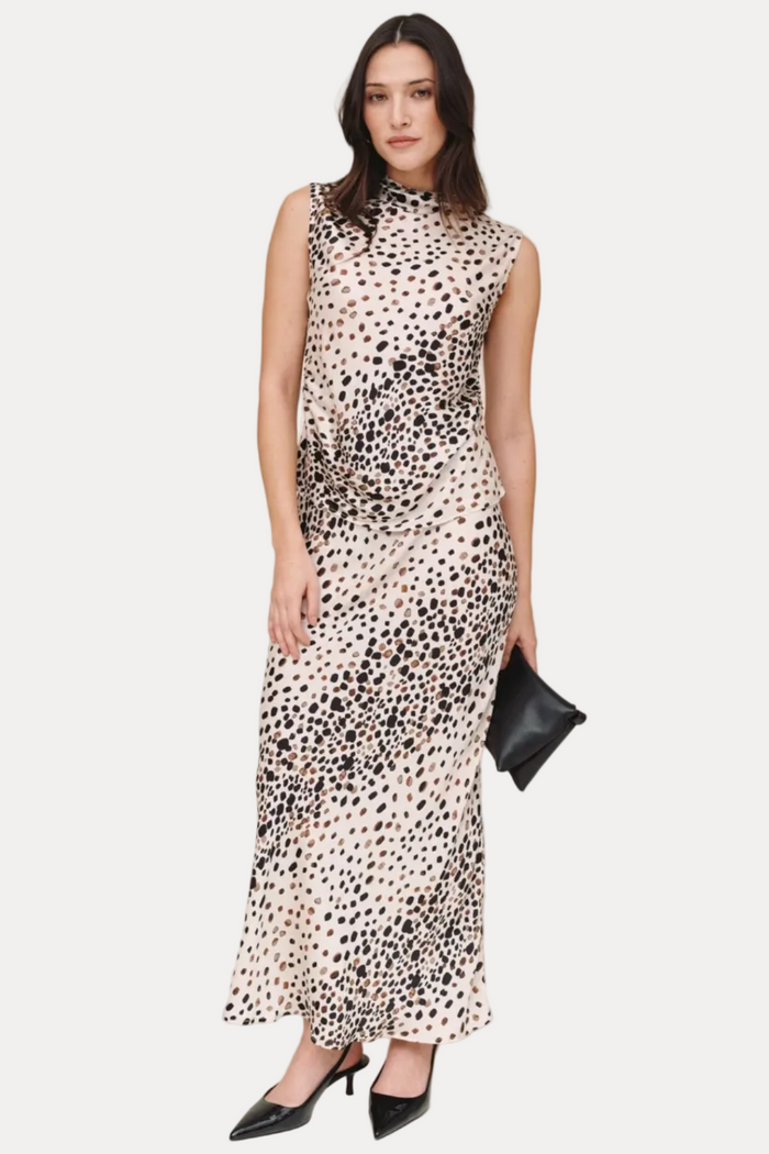 A woman modeling a sleeveless, high-neck maxi dress with a black-on-cream spotted animal print, paired with black pointed-toe heels and holding a small black clutch against a plain beige studio background.