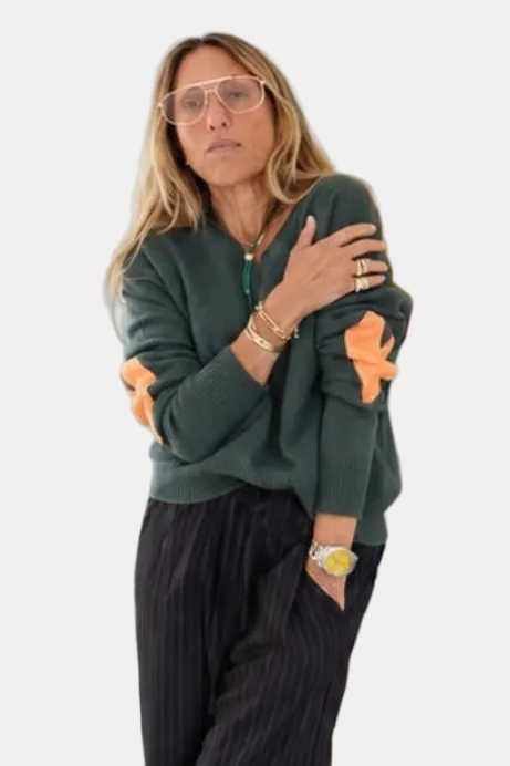 A woman with blonde hair and glasses stands in front of a colorful abstract painting, wearing a dark green knit sweater with orange star details on the sleeves and dark pinstriped trousers.