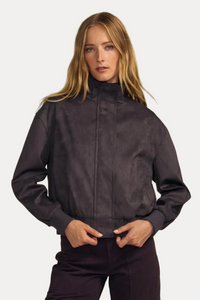 A woman modeling a dark plum zip-up jacket with dark green ribbed cuffs and matching dark plum pants, set against a solid beige studio background.