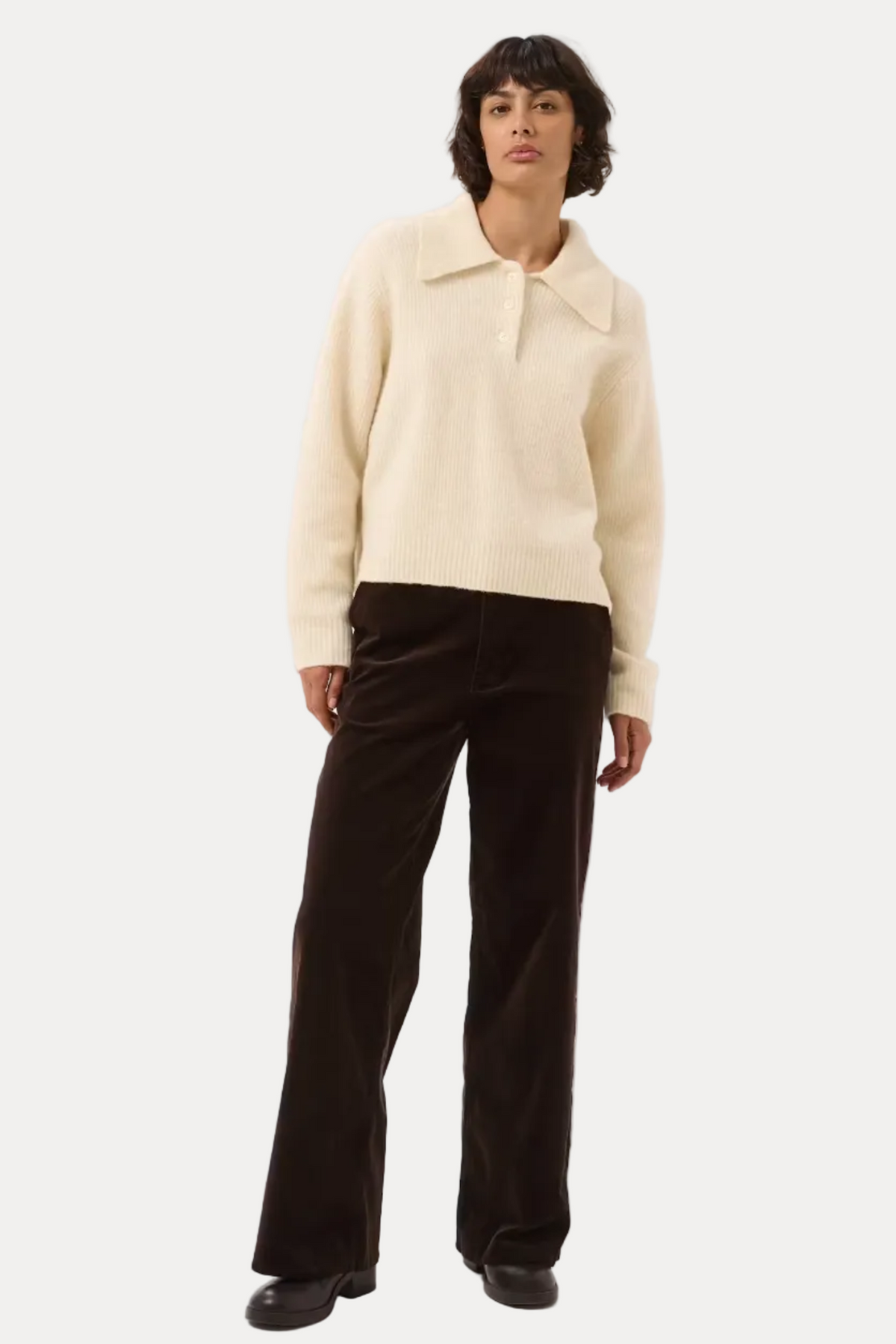 A model stands against a light gray studio background wearing a cream-colored knit polo-collar sweater and dark brown wide-leg trousers.