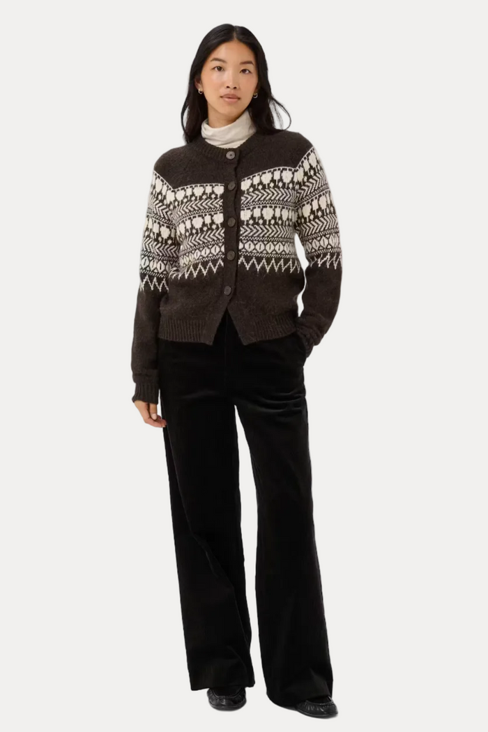 A woman modeling a dark brown cardigan with a cream and brown Fair Isle pattern, paired with black wide-leg trousers and a white turtleneck, against a light gray studio background.