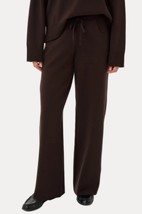 A model wearing dark brown wide-leg trousers with a drawstring waist, paired with a matching dark brown top and black shoes, shown from the chest down on a light gray studio background.