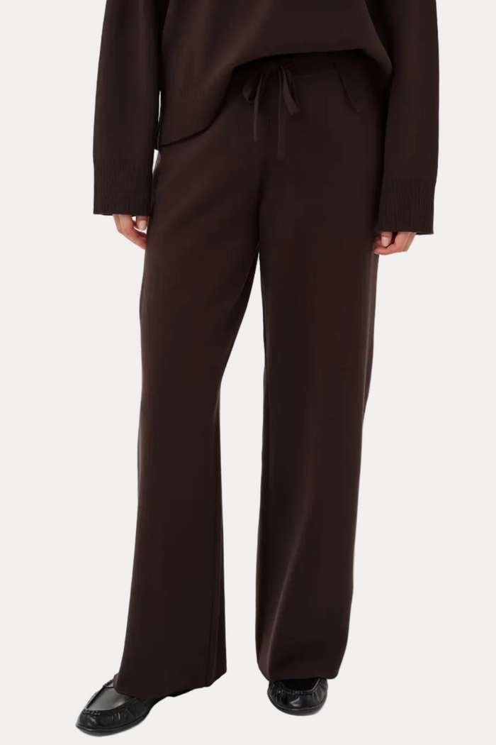 A model wearing dark brown wide-leg trousers with a drawstring waist, paired with a matching dark brown top and black shoes, shown from the chest down on a light gray studio background.