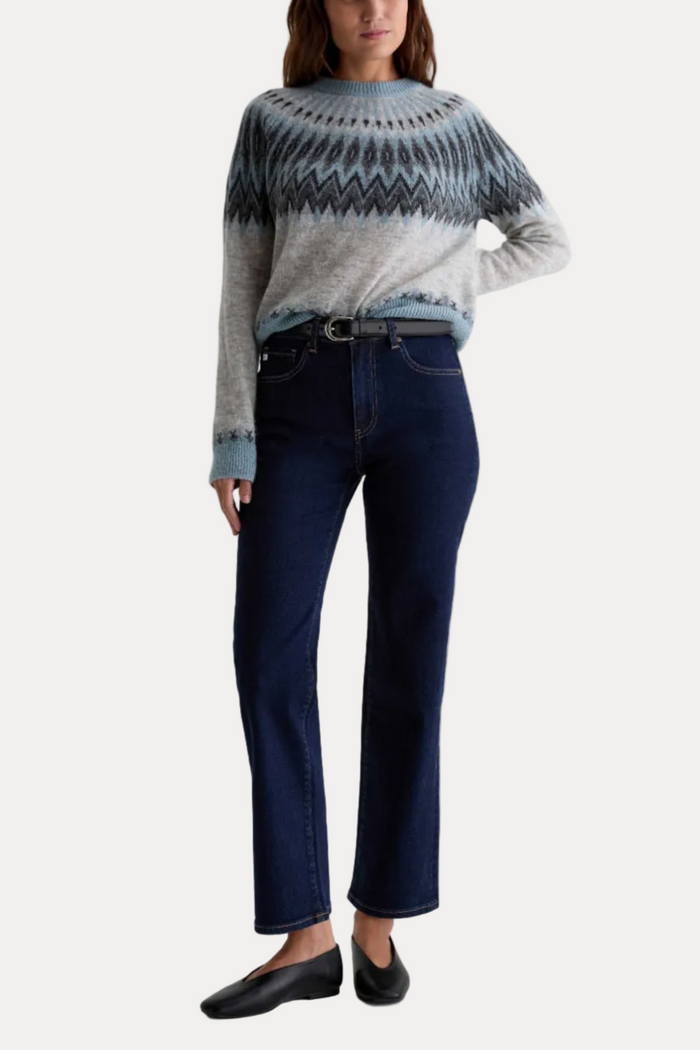 A model wearing a light gray and blue patterned knit sweater, dark blue jeans, and black flats, standing against a plain white studio background.