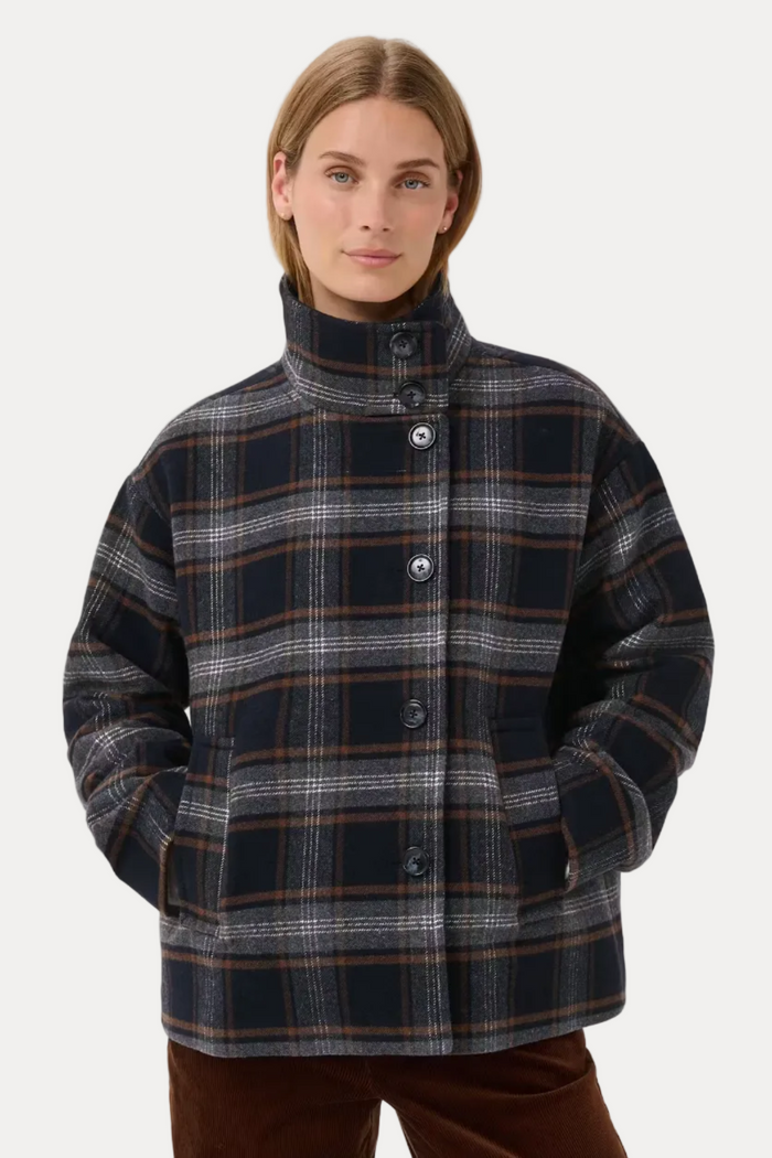 A woman wearing a dark navy and gray plaid button-up jacket with brown and orange stripes, paired with brown trousers, photographed against a light gray studio background.