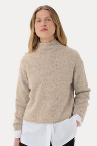 A woman modeling a beige textured knit mock-neck sweater over a white collared shirt and black pants, photographed against a clean white studio background.