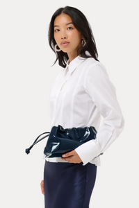 A woman wearing a white long-sleeve button-up shirt and navy blue satin skirt, holding a shiny dark navy drawstring pouch bag around her waist, against a light gray studio background.