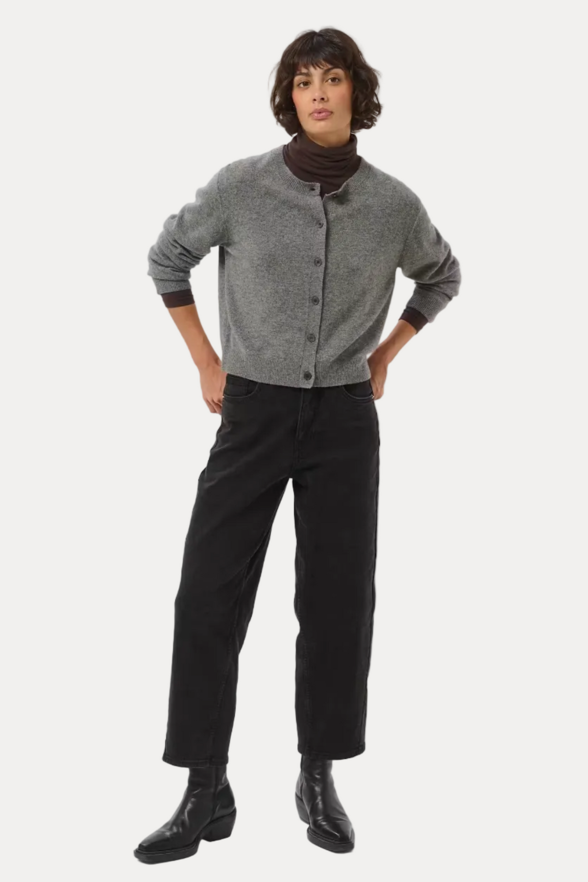 A model stands against a light gray studio background wearing a gray button-front cardigan over a dark brown turtleneck, black wide-leg trousers, and black leather boots.