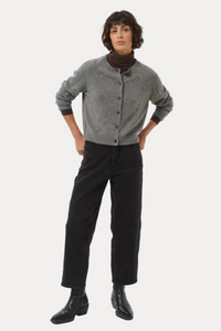 A model stands against a light gray studio background wearing a gray button-front cardigan over a dark brown turtleneck, black wide-leg trousers, and black leather boots.