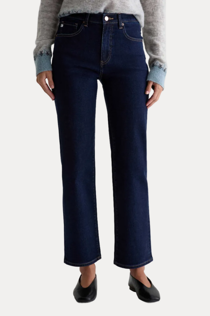 A person wearing dark blue straight-leg denim jeans with a gray knit sweater and black flats, shown from the waist down against a plain light gray studio background.
