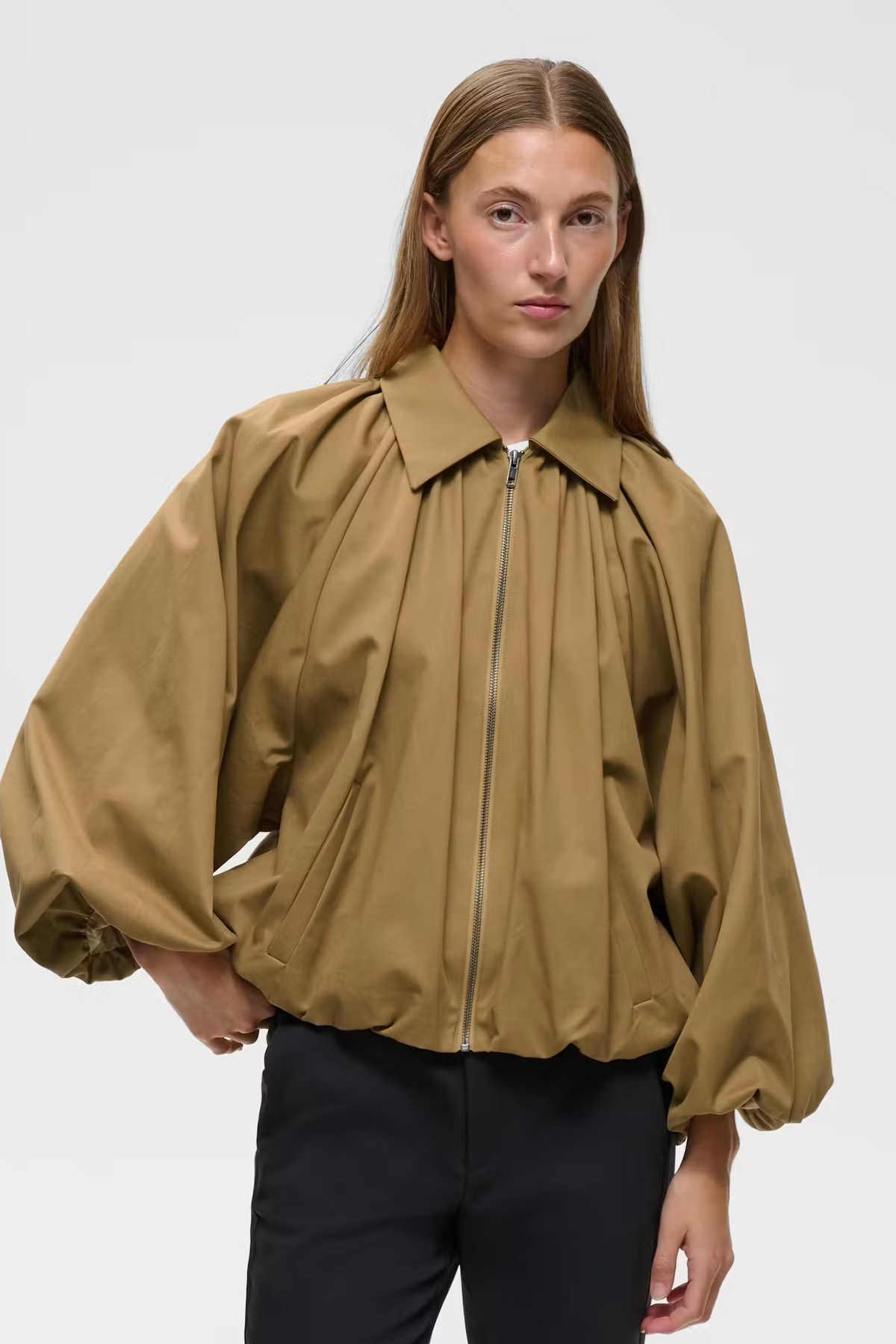 A model wearing a cropped, oversized zip-up jacket in a warm camel brown color with voluminous balloon sleeves, paired with black trousers, against a plain light gray studio background.