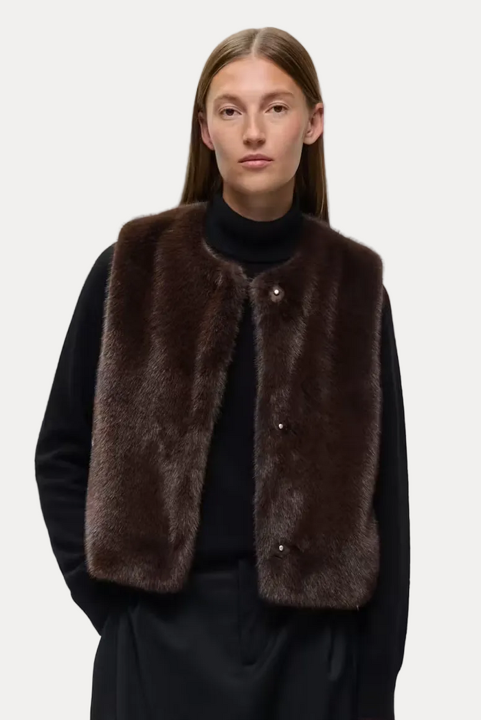 A model stands against a light gray studio background wearing a dark brown faux-fur vest over a black turtleneck and black wide-leg trousers.