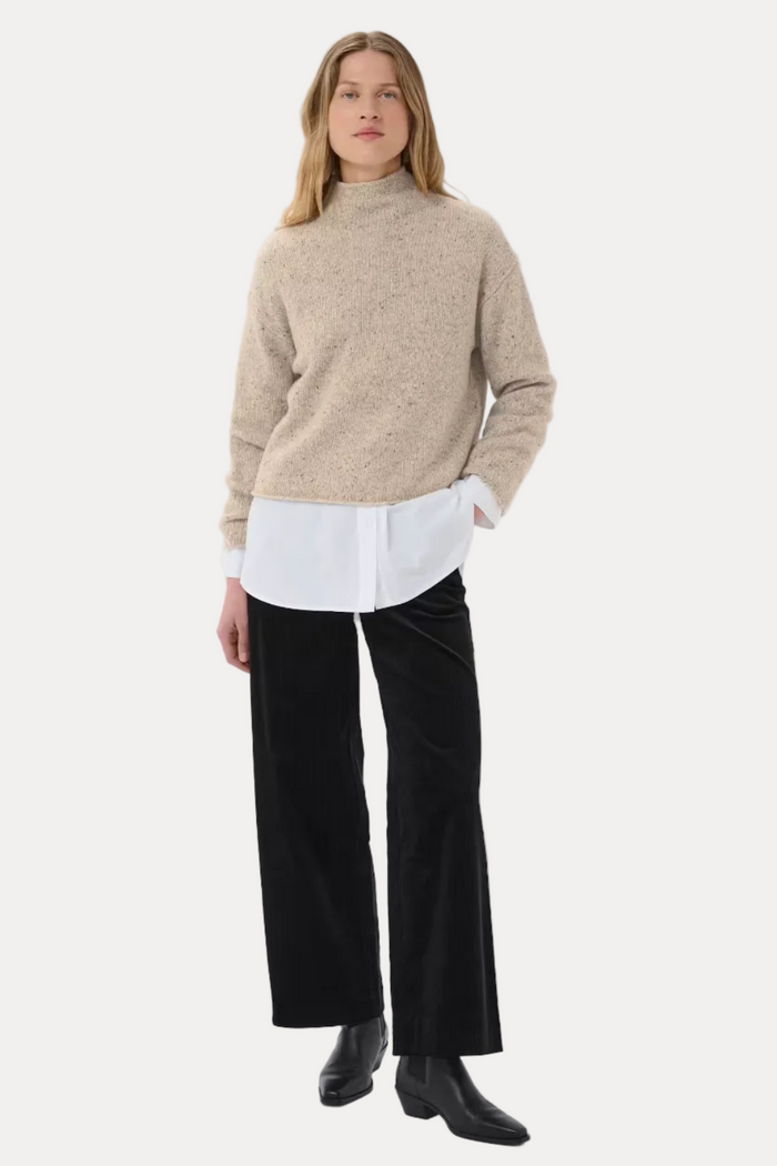 A model stands against a light gray studio background wearing a beige knit mock-neck sweater over a white collared shirt, black wide-leg trousers, and black ankle boots.