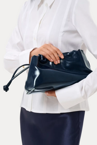 A person in a white button-up shirt and dark navy skirt holds a shiny dark navy clutch bag with a drawstring detail against a clean white studio background.