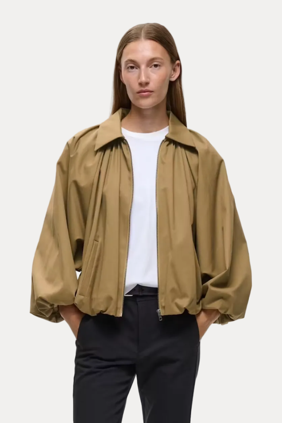 A model stands against a light gray studio background wearing a cropped tan overshirt over a white T-shirt, black cropped trousers, and black loafers.