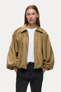 A model stands against a light gray studio background wearing a cropped tan overshirt over a white T-shirt, black cropped trousers, and black loafers.