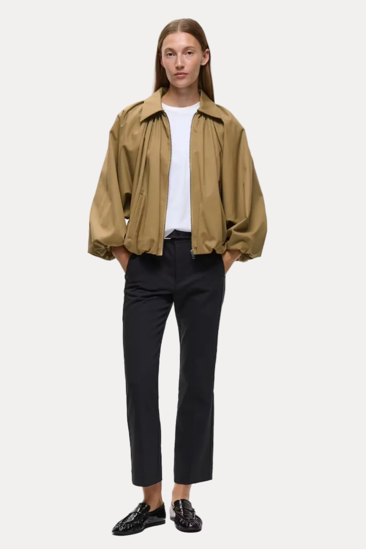A model stands against a light gray studio background wearing a cropped tan overshirt over a white T-shirt, black cropped trousers, and black loafers.