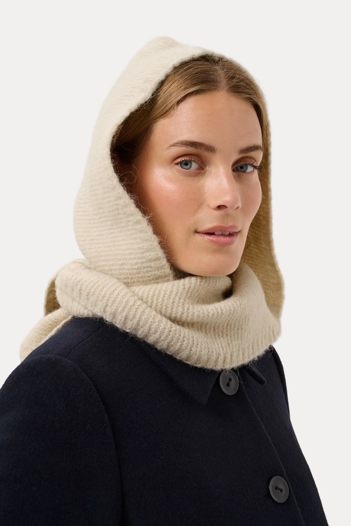 A woman wearing a cream-colored knit hooded scarf or cowl over a dark navy coat, photographed against a plain light gray studio background.