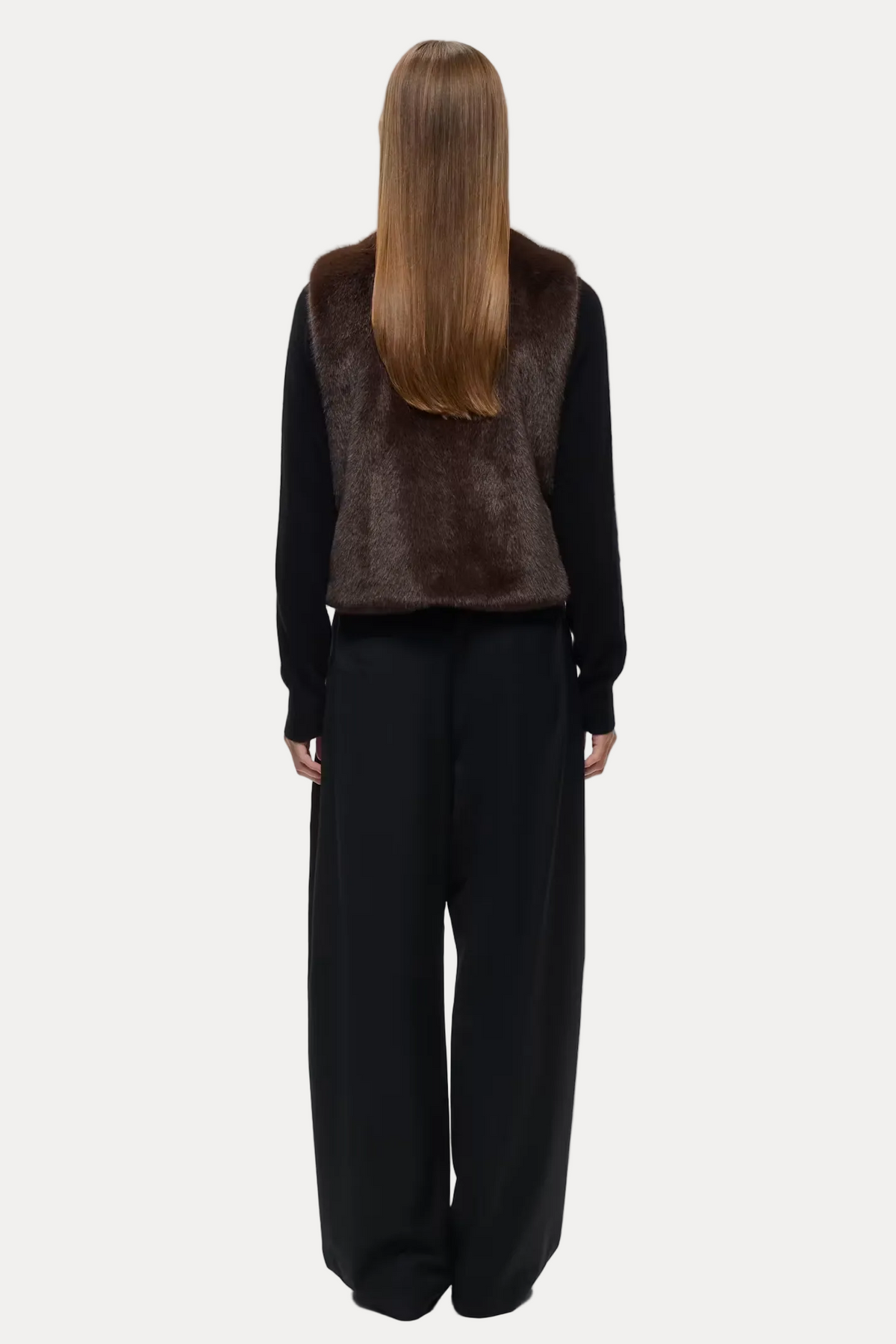 A model shown from the back wearing a dark brown fur vest over a black long-sleeve top and black wide-leg trousers, set against a plain light gray studio background.