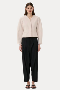 A model stands against a light gray studio background wearing a pale blush cropped wrap jacket, black cropped trousers, and black sandals.