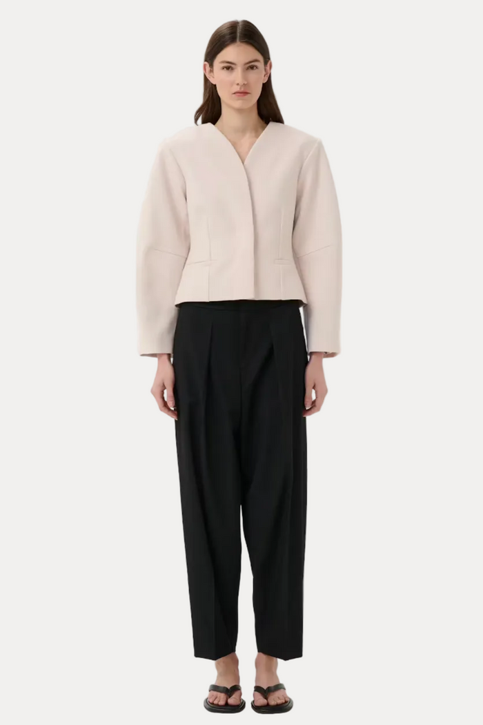 A model stands against a light gray studio background wearing a pale blush cropped wrap jacket, black cropped trousers, and black sandals.