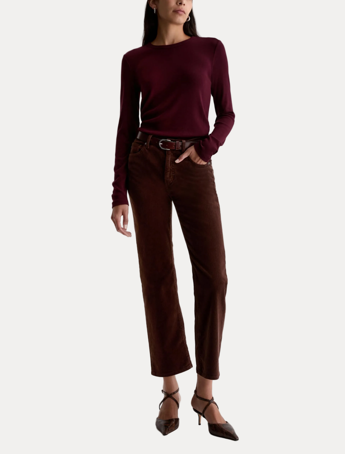 A model wearing a dark burgundy long-sleeve knit top and dark brown cropped trousers, styled with a dark brown belt, brown shoes, and a slim black bag against a plain white studio background.