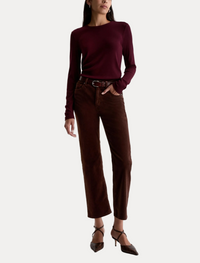 A model wearing a dark burgundy long-sleeve knit top and dark brown cropped trousers, styled with a dark brown belt, brown shoes, and a slim black bag against a plain white studio background.
