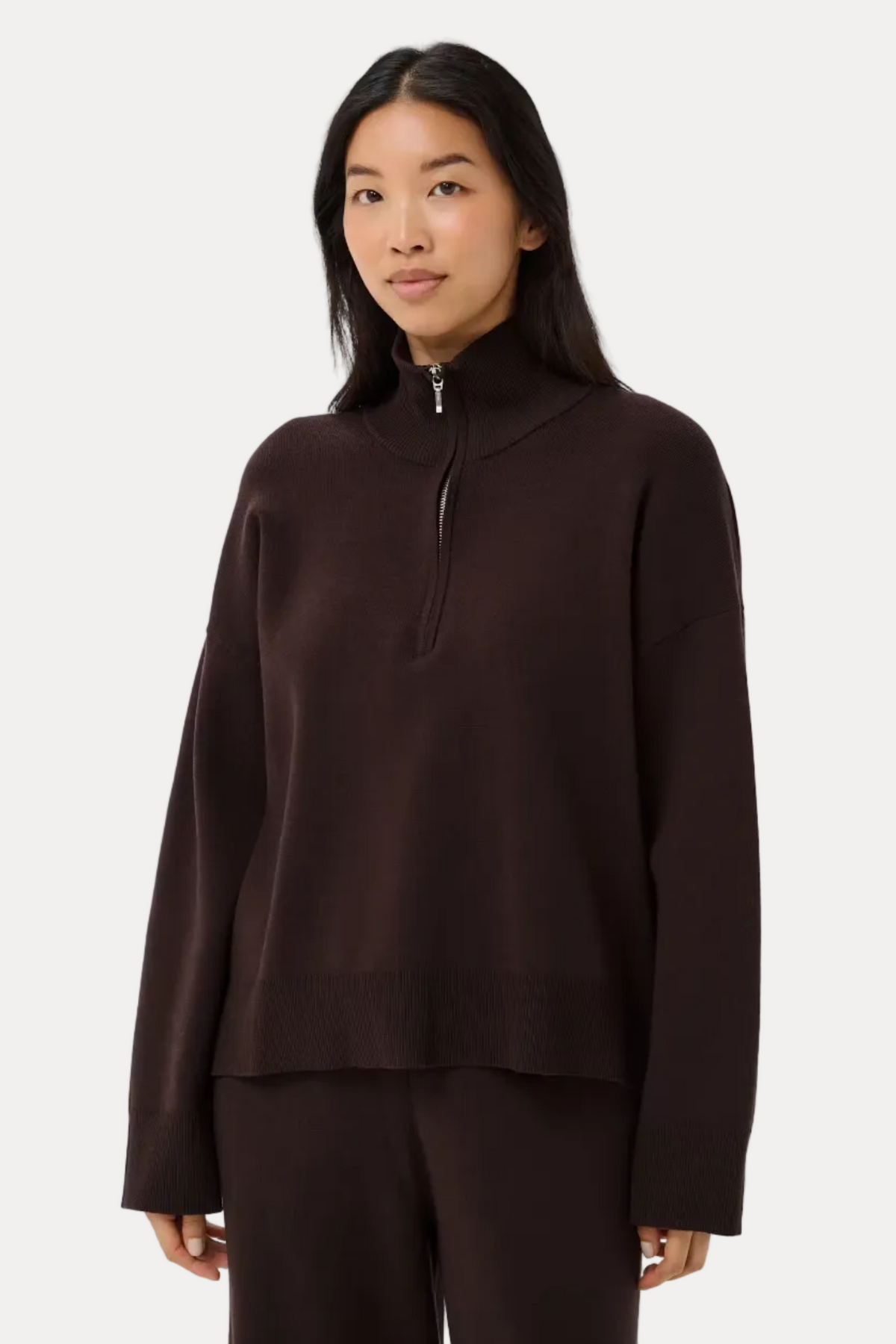 A woman modeling a dark brown long-sleeve quarter-zip pullover and matching dark brown pants against a light gray studio background.