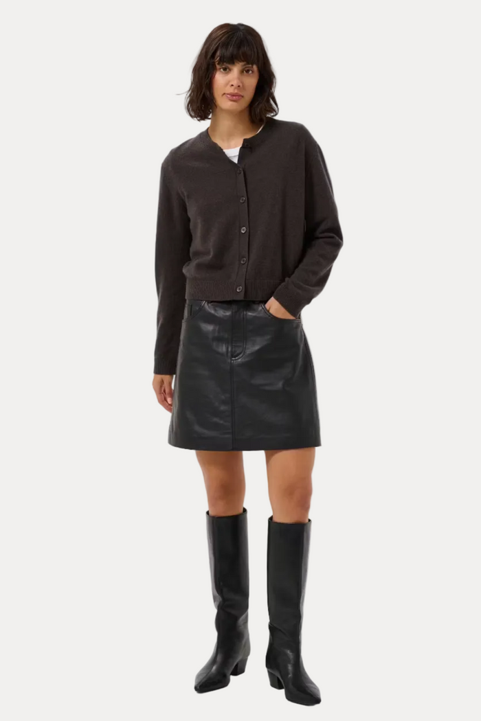 A model stands against a light gray studio background wearing a dark brown cropped button-front cardigan, a black faux-leather mini skirt, and black knee-high boots.