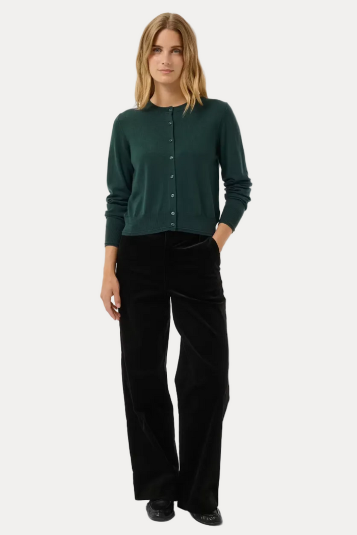 A woman stands against a light gray studio background wearing a dark green long-sleeve button-front cardigan and black wide-leg trousers.