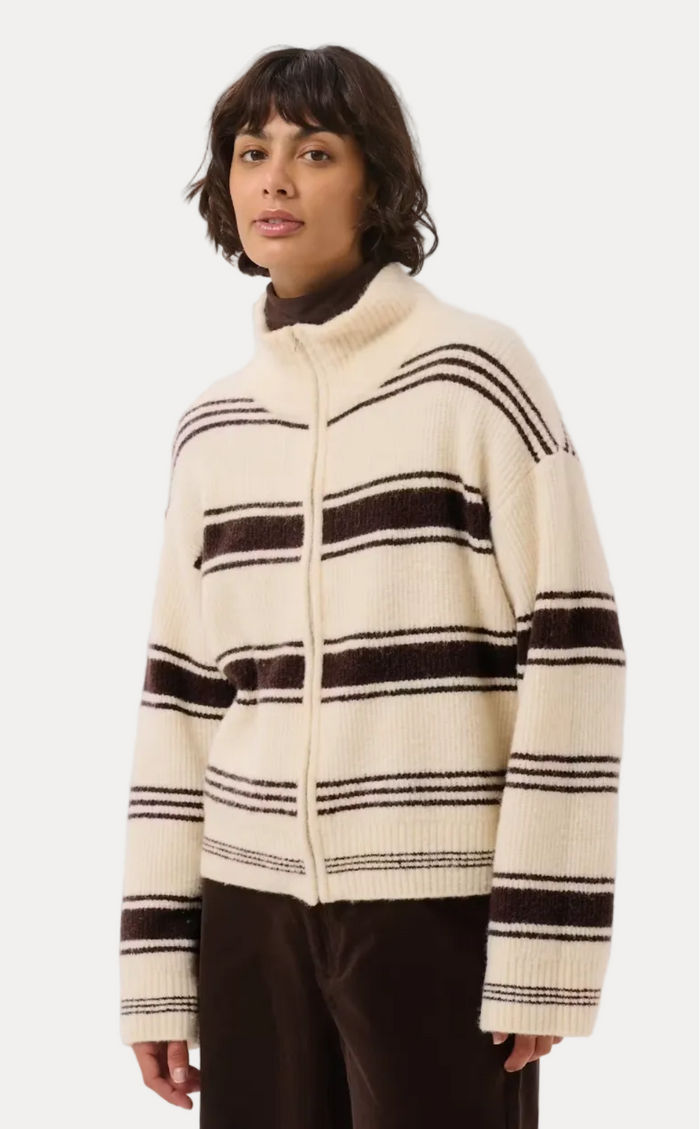 A model wearing a cream-colored knit zip-up cardigan with dark brown horizontal stripes, paired with dark brown trousers, against a light gray studio background.