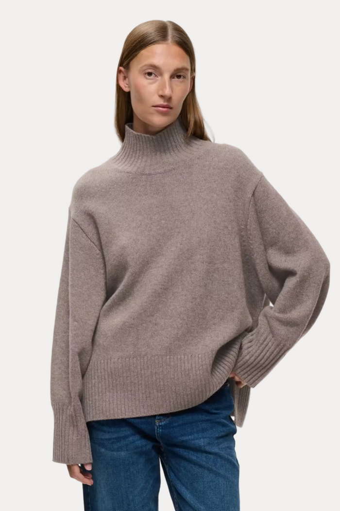 A woman modeling an oversized taupe knit turtleneck sweater with blue jeans against a light gray studio background.