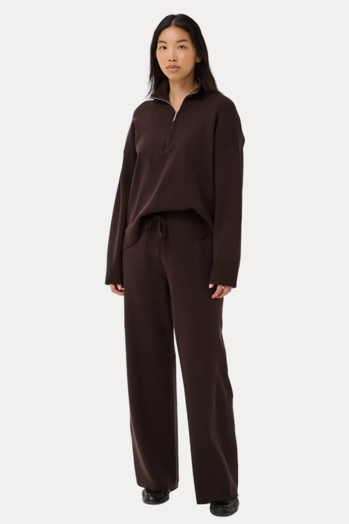 A model stands against a light gray studio background wearing a matching dark brown lounge set consisting of a long-sleeve half-zip pullover and wide-leg drawstring pants.
