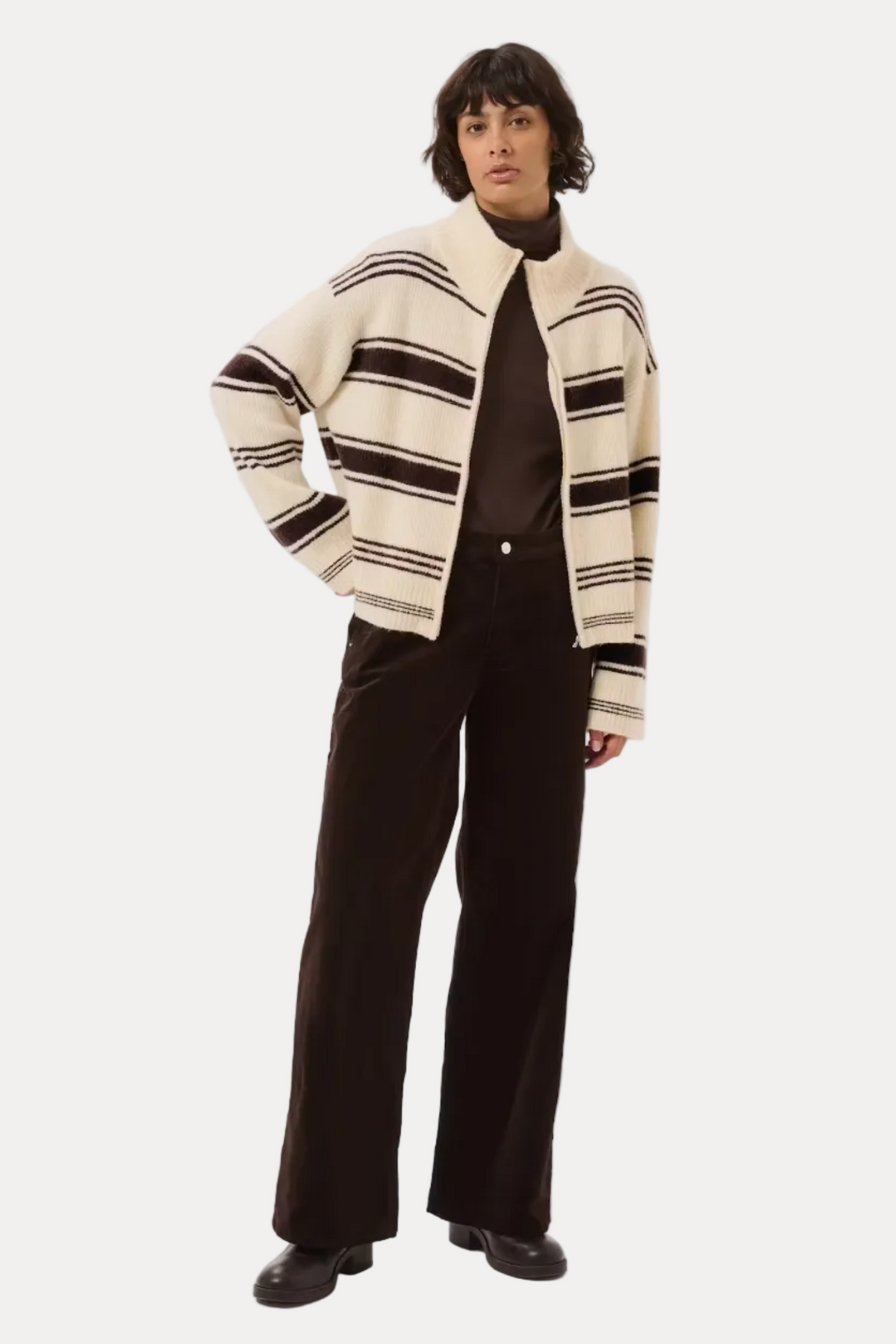 A model stands against a light gray studio background wearing a cream and dark brown striped knit zip-up cardigan over a dark brown turtleneck, paired with dark brown wide-leg trousers and dark brown shoes.