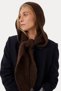 A woman wearing a dark brown fuzzy hooded scarf or head covering tied around her neck, paired with a dark coat, against a plain light gray studio background.