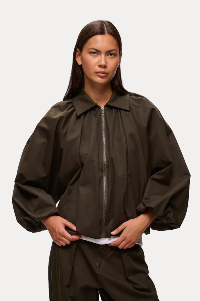 A model wearing a dark brown oversized zip-up jacket with voluminous balloon sleeves and matching dark brown trousers, photographed against a light gray studio background.