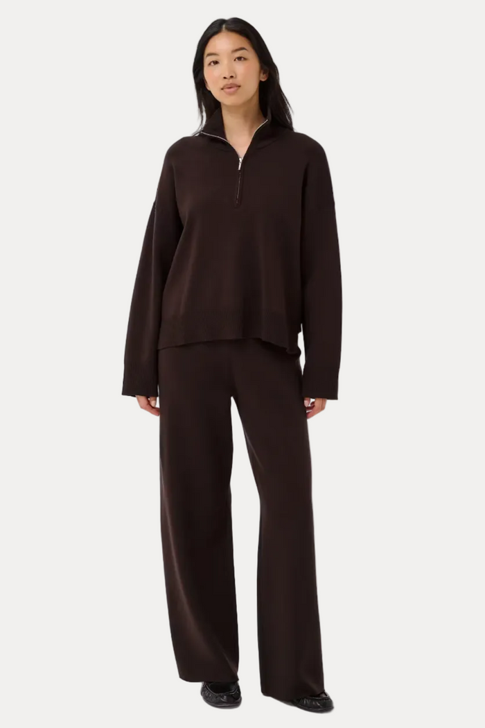 A woman modeling a dark brown two-piece knit loungewear set, consisting of a long-sleeve quarter-zip pullover and matching wide-leg pants, against a plain light gray studio background.