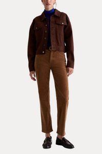 A model is shown from the neck down wearing a dark brown cropped denim jacket, a blue turtleneck, brown corduroy trousers, and dark brown loafers, shot against a plain white studio background.