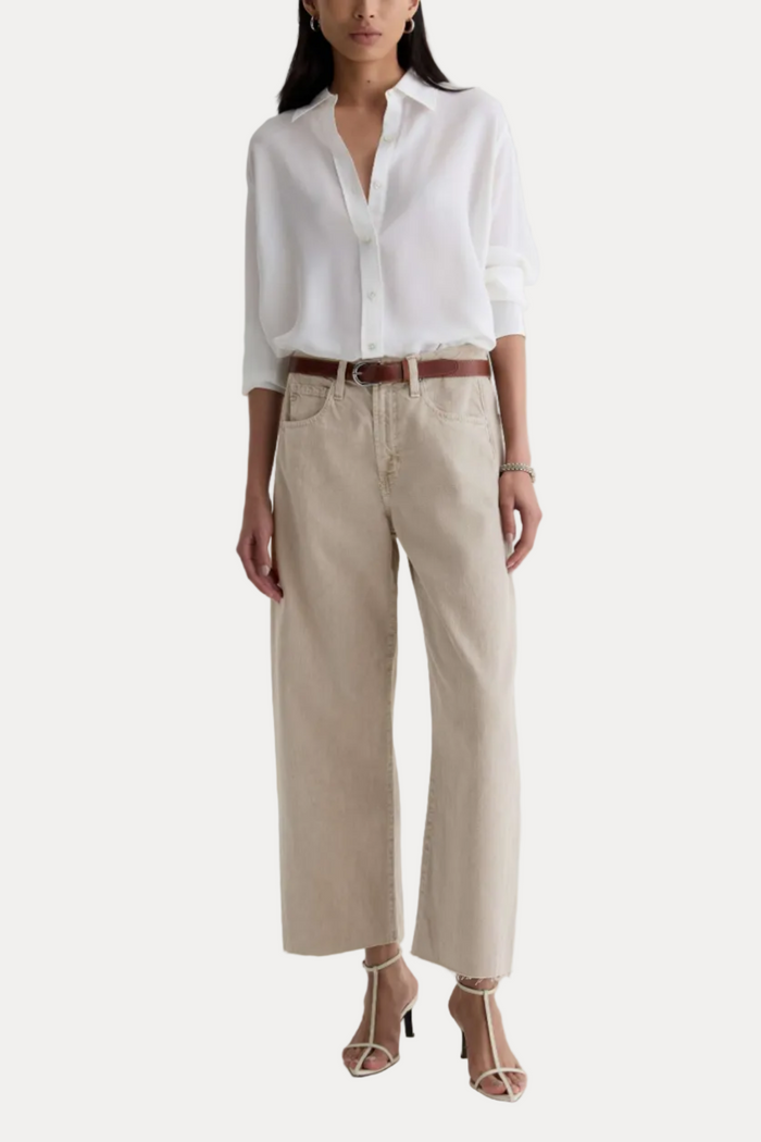 A model wearing a white long-sleeve button-down shirt, beige wide-leg cropped pants, a brown leather belt, and beige strappy high-heeled sandals, standing against a plain off-white studio background.