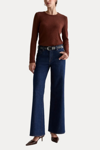 A model wearing a brown ribbed long-sleeve top, dark blue wide-leg jeans with a black belt, and brown pointed-toe heels, standing against a plain white studio background.