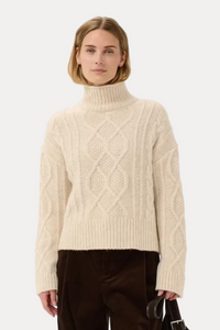 A model stands against a light gray studio background wearing a cream cable-knit turtleneck sweater, dark brown corduroy trousers, and black shoes, while carrying a dark brown tote bag.