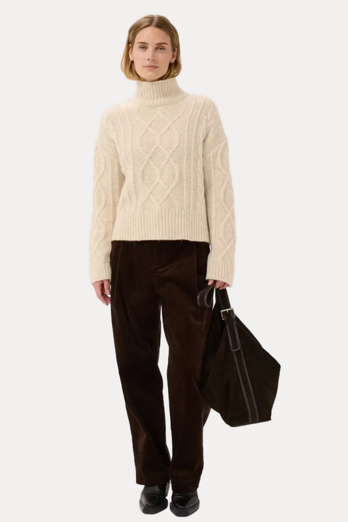 A model stands against a light gray studio background wearing a cream cable-knit turtleneck sweater, dark brown corduroy trousers, and black shoes, while carrying a dark brown tote bag.