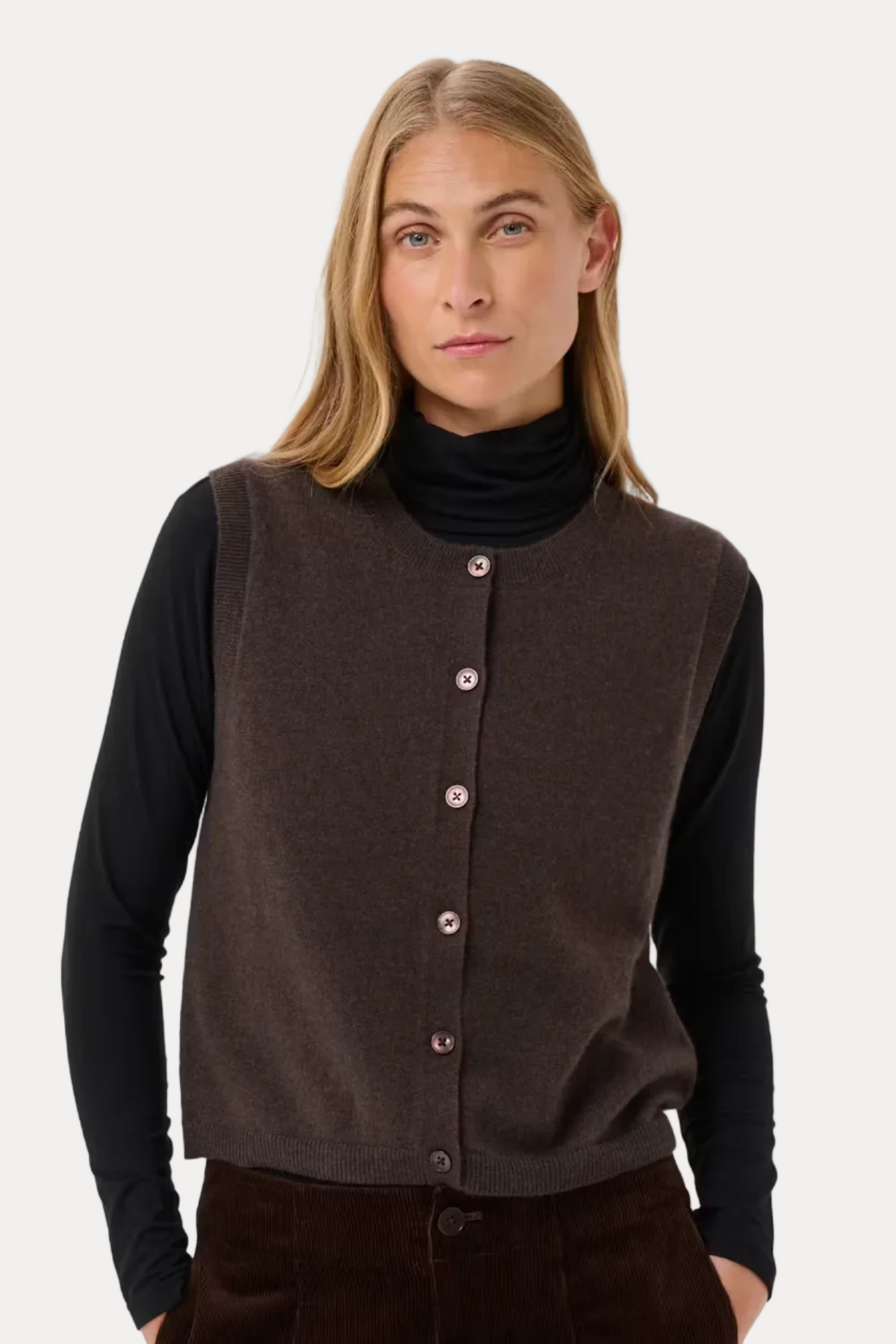 A woman modeling a sleeveless brown knit vest with pink buttons over a black turtleneck and dark brown corduroy pants, shot against a light gray studio background.