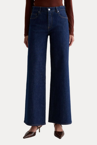 A model wearing dark blue wide-leg denim jeans, a brown long-sleeve top, and brown pointed-toe heels, standing against a plain light gray studio background.
