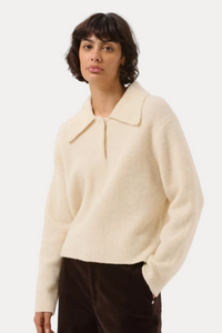 A model wearing a cream-colored ribbed knit polo-collar sweater and dark brown corduroy trousers, standing against a light gray studio background.